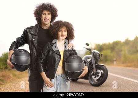 Paar in Lederjacken mit Helmen vor einem geparkten Motorrad auf offener Straße Stockfoto