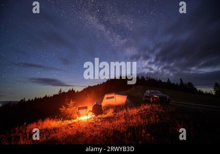 Camping in den Bergen unter dem abendlichen bewölkten Himmel mit Sternen. Rückansicht der Person, die sich am Lagerfeuer ausruhen kann, in der Nähe des Touristenzelts und Geländewagens auf dem Hügel, in der Nacht sitzen und den Sternenhimmel beobachten kann. Stockfoto