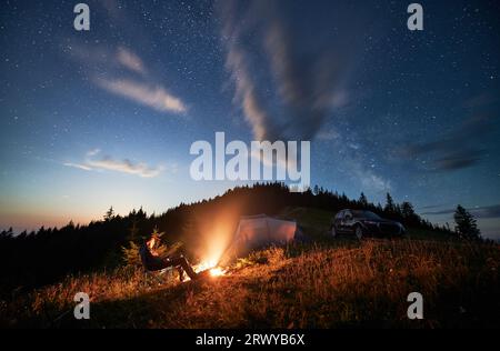 Camping in den Bergen unter bewölktem Nachthimmel. Junger Mann, der sich am Lagerfeuer ausruht, in der Nähe eines Touristenzelts und eines Geländewagens auf einem Hügel. Ein reisender Mann, der nachts die Sterne beobachtet. Stockfoto