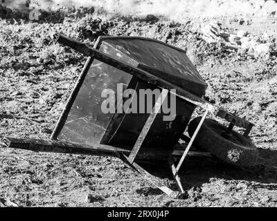 Ein verprügelter alter Schubkarren liegt überfüllt in einem Hinterhof, Werkzeuge liegen auf der Seite im Schmutz, monochrom Stockfoto