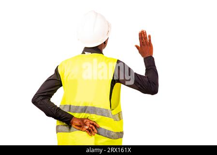 Junger Ingenieur trägt grüne Weste und weißen Schutzhelm, während er mit der Hand hinter dem Rücken Eid leistet. Stockfoto