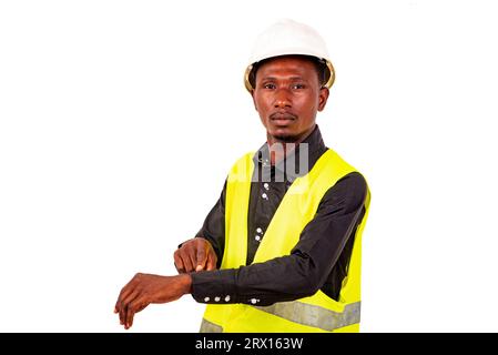 Junger, müder Ingenieur, der grüne Weste und weißen Schutzhelm trägt, während er mit dem Finger auf die Armbanduhr zeigt. Stockfoto