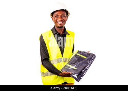 Junger glücklicher Ingenieur, der eine grüne Weste und einen weißen Schutzhelm trägt, während er die Arbeitskleidung hält. Stockfoto