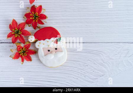Kekse mit dem Gesicht des Weihnachtsmanns auf weißem hölzernem Hintergrund mit roter Blumendekoration. Draufsicht. Leerzeichen kopieren. Stockfoto