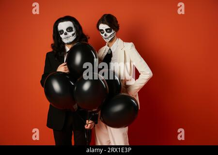 Unheimliches Paar in catrina-Make-up und Anzügen mit schwarzen Ballons auf roter, DIA de los Muertos-Party Stockfoto