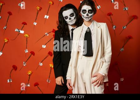 Ehepaar in DIA de los muertos catrina Make-up und Anzügen mit Blick auf die Kamera vor rotem Hintergrund mit Blumen Stockfoto