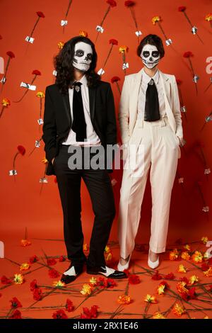 Elegantes Ehepaar in DIA de los muertos-Make-up und Anzügen auf rotem Hintergrund mit Nelkenblüten Stockfoto