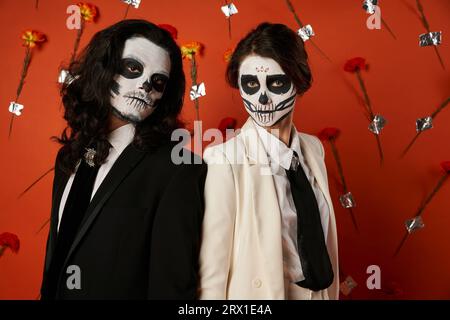 Trendiges Paar in DIA de los muertos Totenkopf-Make-up mit Blick auf die Kamera vor rotem Hintergrund mit Blumen Stockfoto