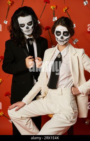 Elegantes Paar in catrina-Make-up und Anzügen auf rotem Hintergrund mit Nelken, Day of Dead Stockfoto