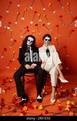 Stilvolles Paar mit Totenkopf-Make-up und Anzügen auf Stühlen auf rotem Hintergrund mit Blumen, Day of Dead Stockfoto