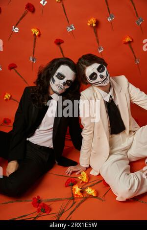 Ein Paar in einem gruseligen catrina-Make-up und Anzügen sitzt auf dem Boden im roten Studio mit Nelkenblüten Stockfoto