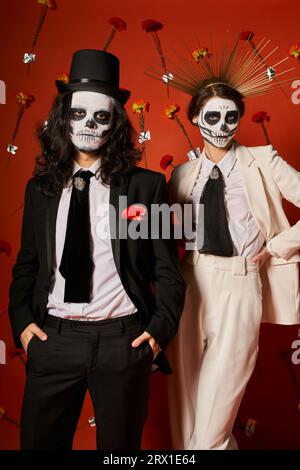 Ehepaar in DIA de los muertos-Make-up und festlichem Outfit mit roter Kulisse und Nelken Stockfoto