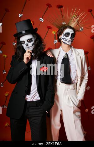 Das Paar trägt ein gruseliges Skelett-Make-up und festliche Kleidung auf rotem Hintergrund mit Blumen, DIA de los muertos Stockfoto