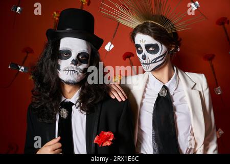 Trendiges Paar in DIA de los muertos Totenkopf-Make-up mit Blick auf die Kamera vor rotem Hintergrund mit Nelken Stockfoto