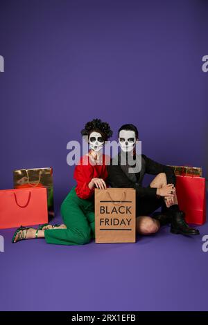 Trendiges Paar in DIA de los muertos Totenkopf-Make-up sitzt in der Nähe von Einkaufstaschen am blauen, schwarzen freitag Stockfoto