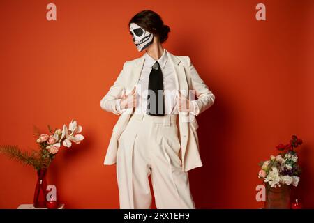 Frau in catrina-Make-up und weißem Anzug in der Nähe des traditionellen Dia de los muertos-Altars mit Blumen auf rot Stockfoto