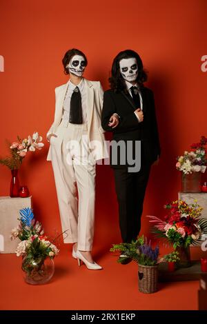 Paar in catrina Make-up und Anzüge in der Nähe von DIA de los muertos ofrenda mit Blumen auf rot Stockfoto