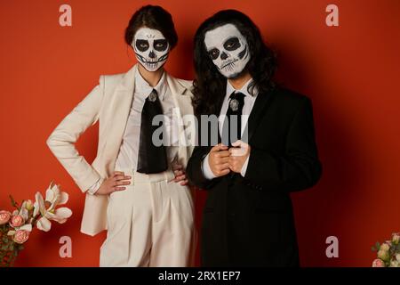 Elegantes Paar in catrina Calavera Make-up mit Blick auf Kamera in der Nähe von Blumendekor auf rot, Day of Dead Stockfoto