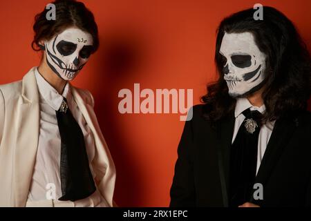 Gruseliges Paar in catrina-Make-up und eleganten Anzügen mit Krawatten auf rot, DIA de los muertos-Tradition Stockfoto