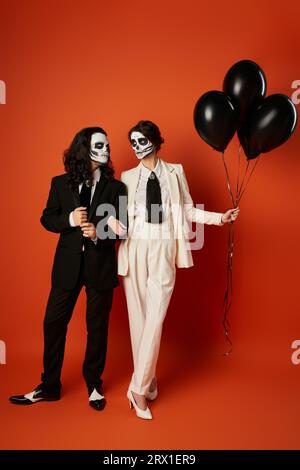 Ein Paar in catrina Make-up und Anzügen, die sich in der Nähe von schwarzen Ballons auf rot, Day of Dead, anschauen Stockfoto