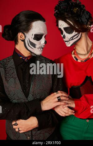 Trendiges Paar im Sugar Skull Make-up, das auf dem roten Dia de los muertos Festival die Hände aneinander berührt Stockfoto