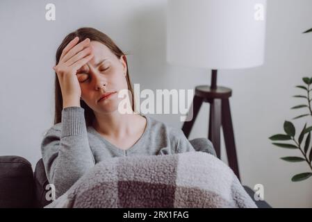 Kranke unglückliche brünette junge Frau bedeckt in Decke sitzend auf Couch im Wohnzimmer zu Hause, den Kopf berührend, leidet unter Kopfschmerzen während Coronaviru Stockfoto