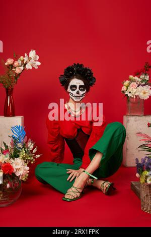 Frau in catrina Make-up sitzt in der Nähe der traditionellen Dia de los muertos ofrenda mit Blumen auf rot Stockfoto