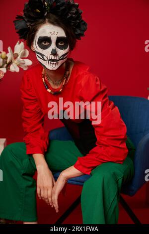 Frau in DIA de los muertos Make-up sitzt im Sessel in der Nähe von Blumen und schaut auf Kamera auf rot Stockfoto