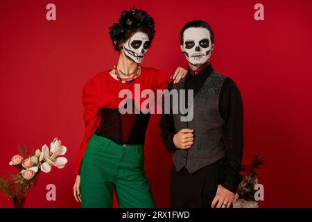 Das stylische DIA de los Muertos-Paar im Totenschädel-Make-up schaut auf die Kamera in der Nähe von Blumen auf rot Stockfoto