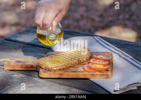 Frau gießt natives Olivenöl auf einen Toast Brot mit frischen Tomaten auf einem Holztisch Stockfoto