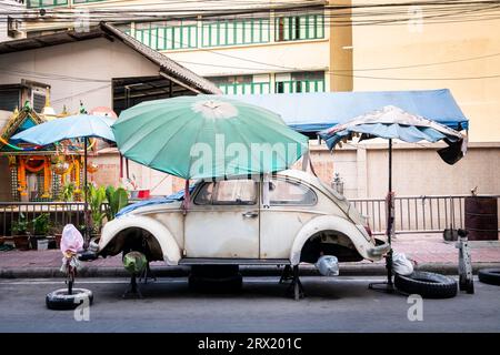 Ein sehr alter, ruinierter Volkswagen Beetle-Wagen sitzt unter ein paar Sonnenschirmen im China Town Viertel von Bangkok, Thailand. Stockfoto