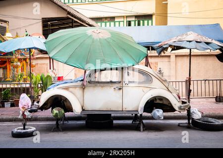Ein sehr alter, ruinierter Volkswagen Beetle-Wagen sitzt unter ein paar Sonnenschirmen im China Town Viertel von Bangkok, Thailand. Stockfoto