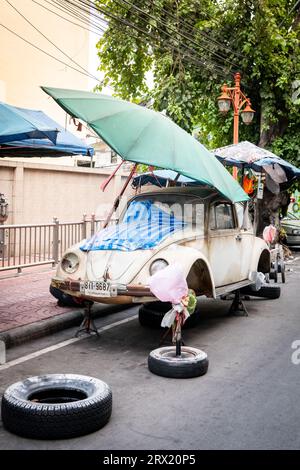 Ein sehr alter, ruinierter Volkswagen Beetle-Wagen sitzt unter ein paar Sonnenschirmen im China Town Viertel von Bangkok, Thailand. Stockfoto