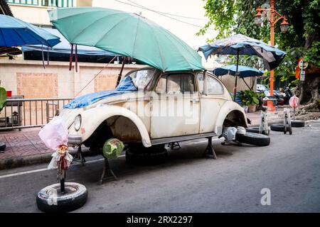 Ein sehr alter, ruinierter Volkswagen Beetle-Wagen sitzt unter ein paar Sonnenschirmen im China Town Viertel von Bangkok, Thailand. Stockfoto