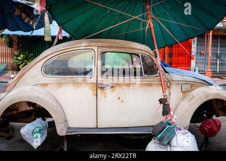 Ein sehr alter, ruinierter Volkswagen Beetle-Wagen sitzt unter ein paar Sonnenschirmen im China Town Viertel von Bangkok, Thailand. Stockfoto