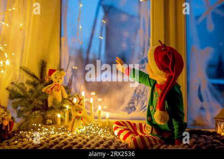 Niedliches Kleinkind in Pyjama, das auf einer wütsamen Decke neben einem Fenster sitzt und draußen auf ein Feuerwerk am Silvesterabend blickt Stockfoto