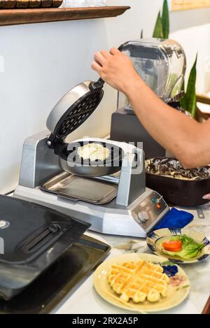 Der Koch gibt den Teig für süße Waffeln mit einem Löffel in das Waffeleisen Stockfoto