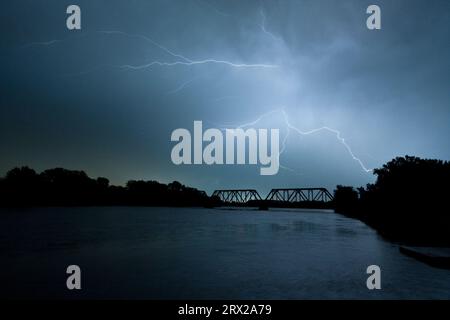 Blitze treffen mit unheimlich schönen Gabeln durch einen gefährlichen grauen Nachthimmel mit einer eisernen Truss-Eisenbahnbrücke über den des Moines River. Stockfoto