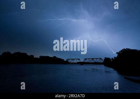 Blitze treffen mit unheimlich schönen Gabeln durch einen gefährlichen, lapisblauen Nachthimmel mit einer eisernen Truss-Eisenbahnbrücke über den des Moines River. Stockfoto