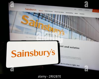 Person, die ein Mobiltelefon mit dem Logo des britischen Supermarktunternehmens J Sainsbury plc auf dem Bildschirm vor der Geschäftswebseite hält. Konzentrieren Sie sich auf das Display des Telefons. Stockfoto