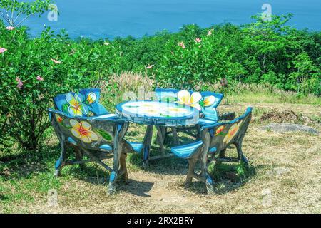Hölzerne bemalte Picknickstühle und runder Tisch im Sommer Hinterhof. Essholz, leere bunte Möbel, grüne Blumenbüsche und blaues Meer bei Sonnenschein Stockfoto