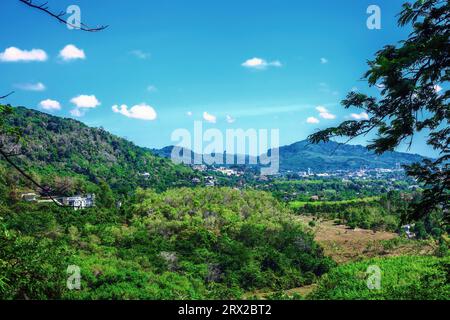 Sommerlandschaft aus tropischem Wald und Stadt in der Ferne in Thailand. Blick aus der Vogelperspektive auf den grünen Regenwald und die Hügel am blauen Himmel Stockfoto