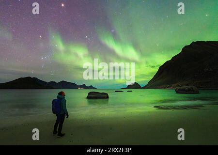 Mann mit Rucksack, der die hellgrünen Lichter der Aurora Borealis (Nordlichter) vom Haukland Beach, den Lofoten-Inseln, Nordland, Norwegen aus bewundert Stockfoto