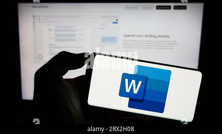 Person, die ein Mobiltelefon mit dem Logo der Textverarbeitungssoftware Microsoft Word auf dem Bildschirm vor der Geschäftswebseite hält. Konzentrieren Sie sich auf das Display des Telefons. Stockfoto