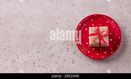 Rote Platte mit weißen Punkten und einem Geschenk aus Bastelpapier, gebunden mit einem roten Band auf einem hellbeigen Hintergrund. Einfacher weihnachtshintergrund oder -Tisch. Stockfoto