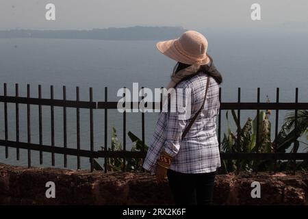 Blick von hinten auf die junge Frau, die das Meer von der umzäunten Terrasse aus bewundert. Stilvolle weibliche Reisende im Sommer Hut mit Blick auf das schöne Meer während des Urlaubs Stockfoto