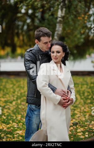 Ein Mann umarmt seine schwangere Frau. Ein Mann in einer schwarzen Jacke umarmt eine Frau in einem weißen Mantel Stockfoto