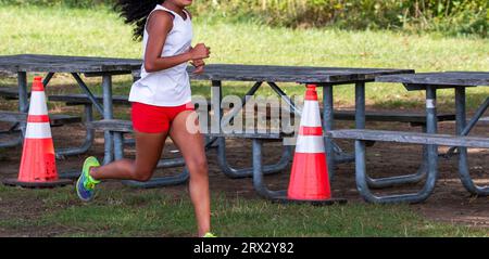 Seitenansicht eines Highschool-Mädchens, das bei einem 5K-Cross County-Rennen neben einem Picknicktisch läuft. Stockfoto