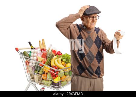 Älterer Mann mit einem Einkaufswagen, der eine Rechnung anschaut und seinen Kopf ungläubig isoliert auf weißem Hintergrund hält Stockfoto