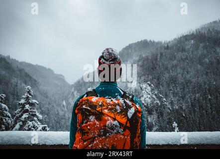 Man Tourist und Kletterer mit orangem Rucksack, der am Aussichtspunkt (Ort) steht, mit fantastischem Blick auf verschneite Berge im kalten Winter. Backpacker Stockfoto